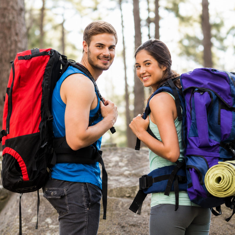 A man and a woman hiking in a forest, each carrying a large backpack. The man wears a red and black backpack and a blue tank top, while the woman wears a purple and blue backpack and a light green tank top.