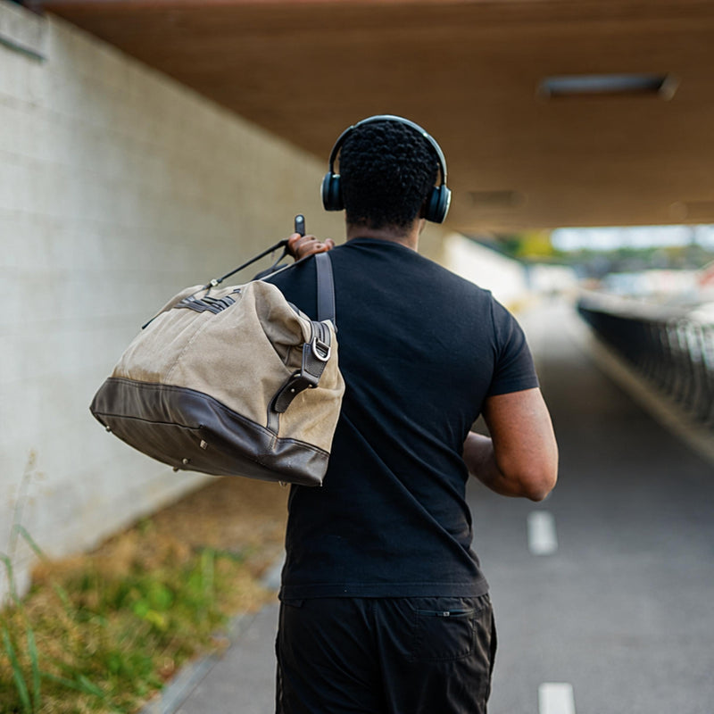 A man seen from behind walking under a concrete bridge, wearing a black t-shirt, black shorts, and over-ear headphones while carrying a beige and dark brown canvas duffel bag.