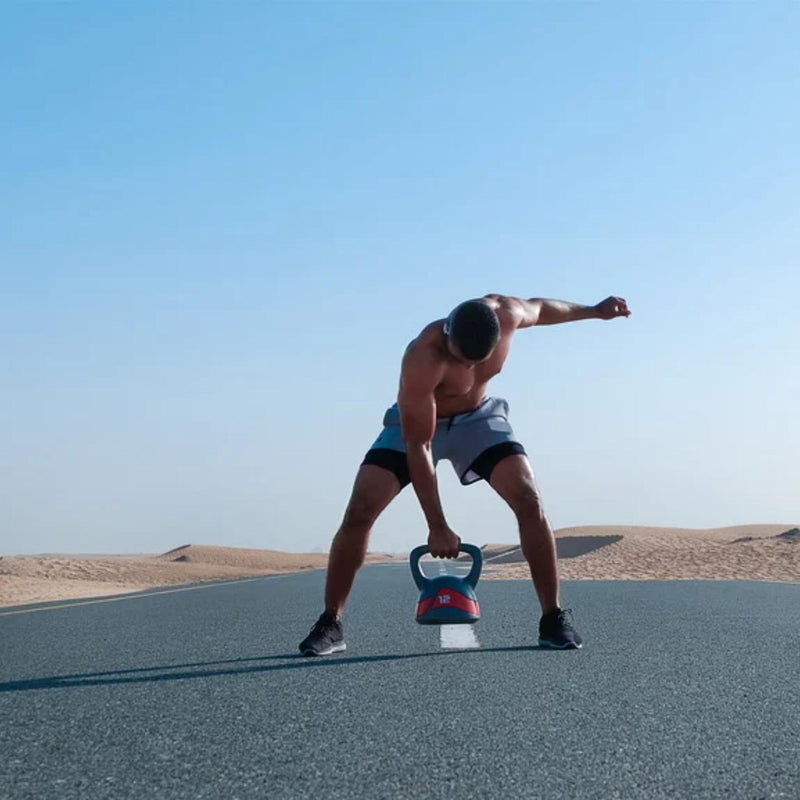 A shirtless man in gray shorts exercises with a kettlebell on an open paved road under a clear blue sky.