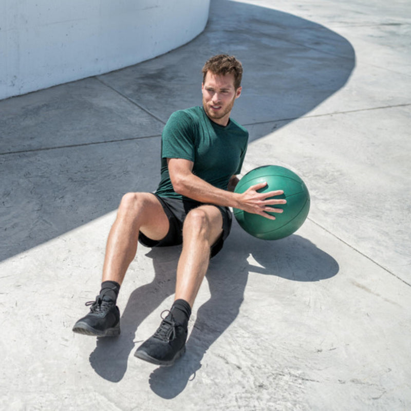 A man in a green t-shirt and black shorts sits on a concrete outdoor court holding a green medicine ball.