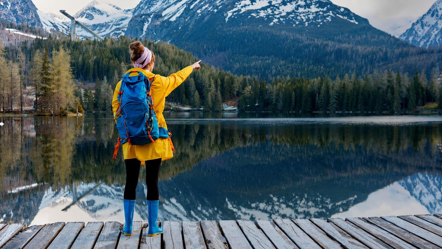 A woman stands on a wooden dock by a calm mountain lake, wearing a bright yellow jacket, blue backpack, and blue rain boots while pointing toward the snowy peaks and forested shoreline.
