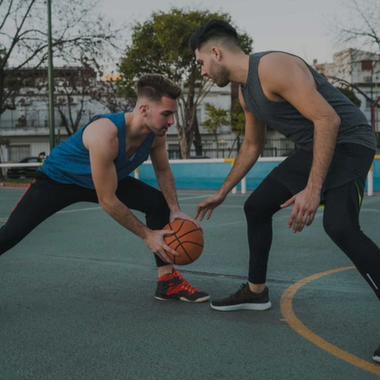 Two men playing basketball on an outdoor court, one in a blue sleeveless top and black pants dribbling the ball while the other in a dark gray sleeveless top and black pants defends him.