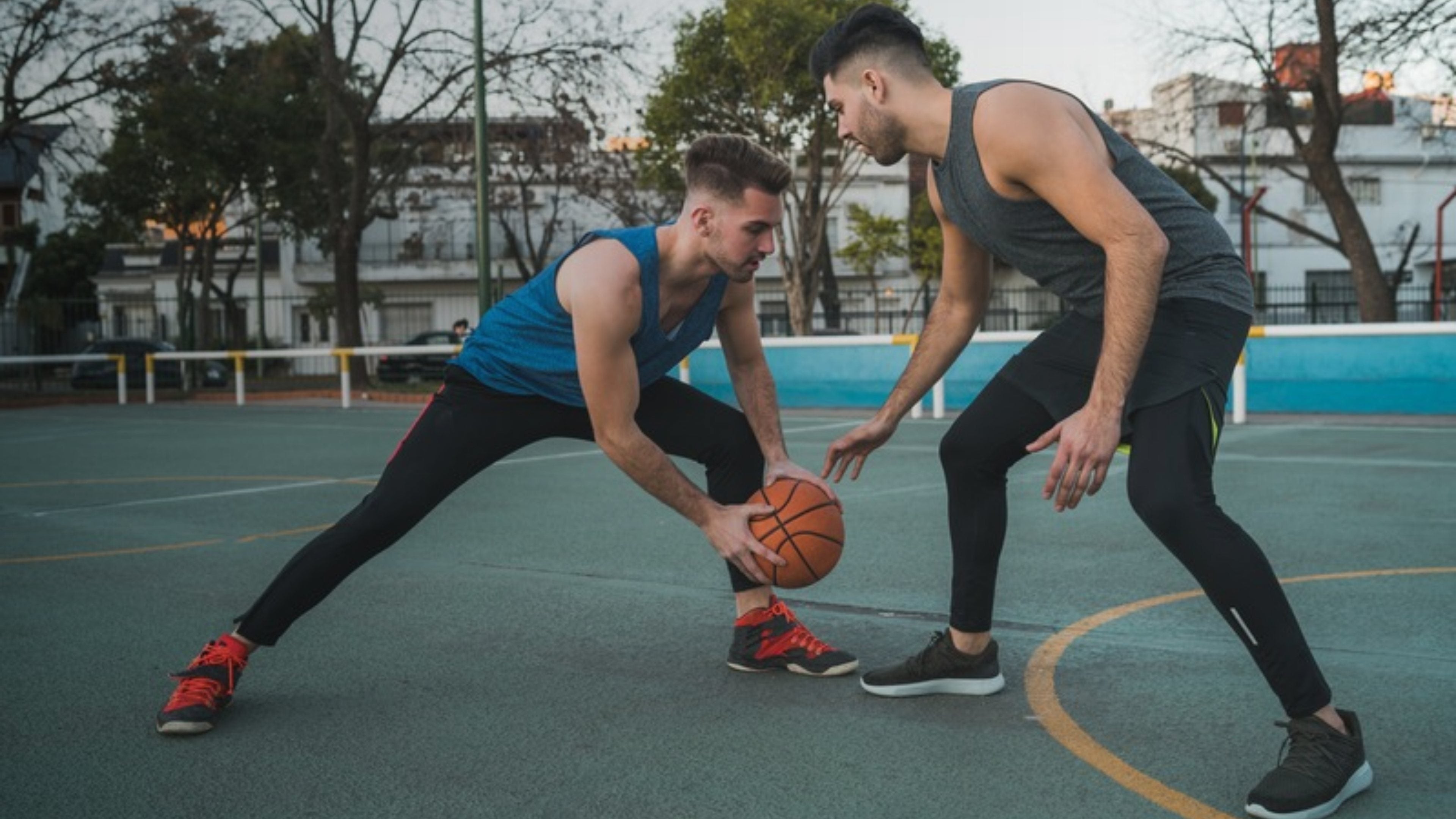 Two men playing basketball on an outdoor court, one in a blue sleeveless top and black pants dribbling the ball while the other in a dark gray sleeveless top and black pants defends him.