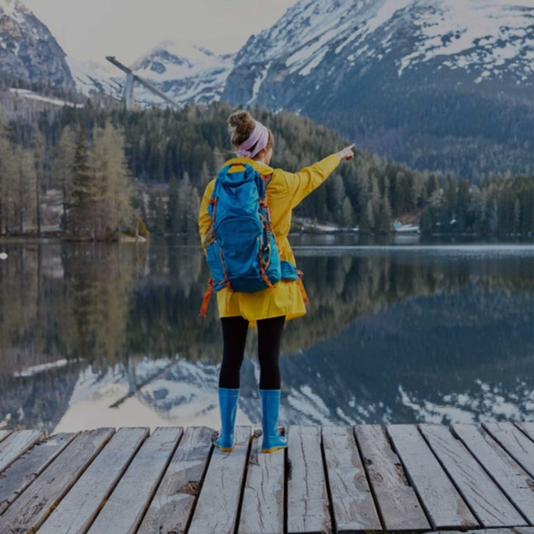 A woman stands on a wooden dock by a calm mountain lake, wearing a bright yellow jacket, blue backpack, and blue rain boots while pointing toward the snowy peaks and forested shoreline.